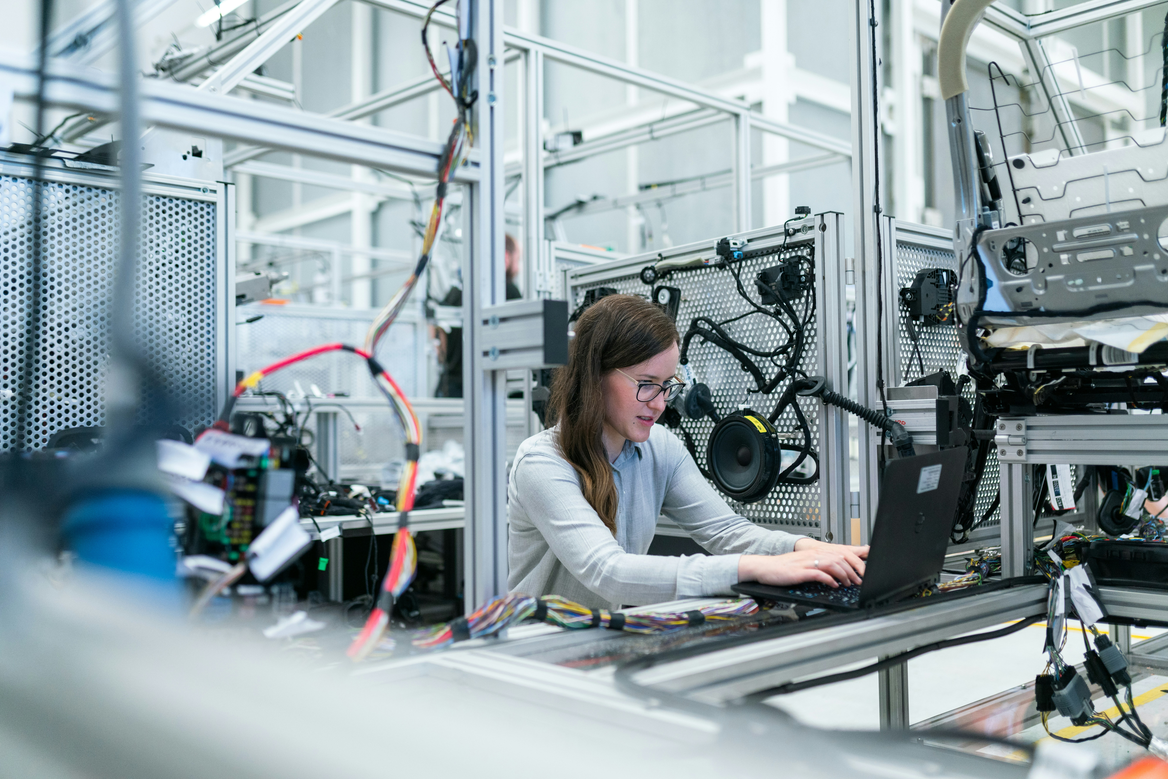 Engenheira trabalhando em um laboratório industrial, usando laptop em meio a equipamentos e cabos de automação.